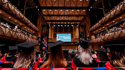 Four women sit in a large auditorium in a university graduation ceremony with their back to the camera wearing a mortar board and gown