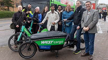 A group of nine people stood behind a green cargo bike and a green electric bike, with a bus at a bus stop in the background