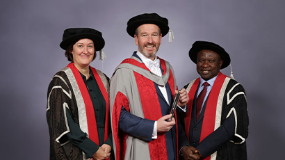 Three people stand together smiling at the camera wearing graduation robes.