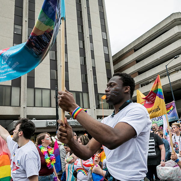 A man taking part in a Pride march and waving a large pride flag, smiling.