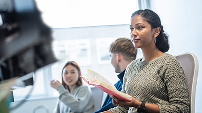 Student sat at a desk looking at a computer screen in the library while two students talk behind her.